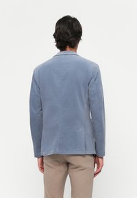 Man seen from behind wearing a light blue blazer and beige trousers against a plain white background.