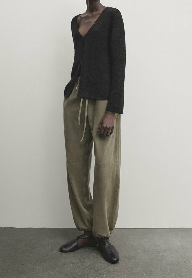 Personne à la peau foncée portant un cardigan noir à col en V profond, un pantalon ample vert olive avec cordon, et des chaussures noires à bout fendu, debout sur un sol gris.