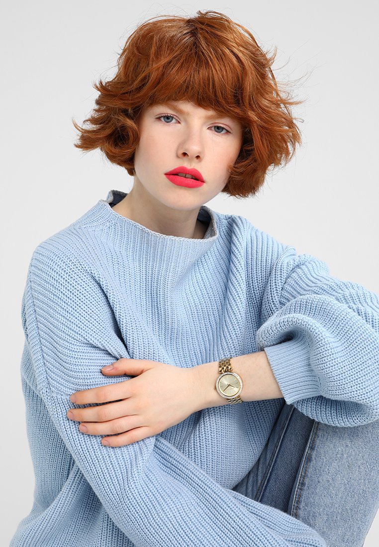 Persona con capelli rossi e ricci di lunghezza corta, che indossa un maglione di lana azzurro chiaro e un orologio dorato al polso, seduta con le braccia incrociate sulle ginocchia.