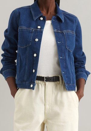 Dark blue denim jacket with silver buttons over white ribbed top, paired with high-waisted beige pants and black belt, hands in pockets.