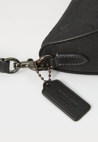Schwarze Stoffhandtasche mit strukturiertem Muster, Lederakzenten, Metallbeschlägen und einem angehängten Etikett, das mit einer Kette befestigt ist.