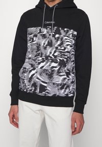Sudadera negra con capucha que presenta un diseño gráfico blanco con patrones abstractos. El diseño incluye un logotipo en la parte superior central. Material de mezcla de algodón.