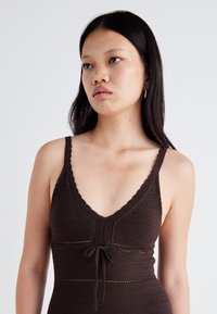 Ruda megzta suknelė su V formos iškirpte, banguotais petnešomis ir juosmens juosta. Yra tekstūruota siuvinėjimo ir horizontalūs raštai.