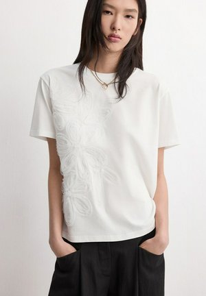 Giovane con capelli lunghi e scuri indossa una maglietta bianca a maniche corte con una trama floreale su un lato e pantaloni neri, con le mani nelle tasche.