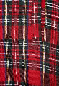 Czerwona tkanina w kratę z wzorem w kratkę, zawierająca czarne, zielone, białe i żółte paski. Materiał ma teksturowany wygląd tkaniny.