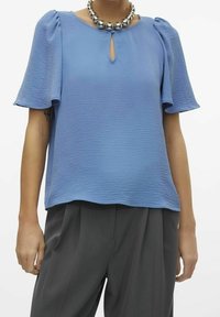 Blusa azzurro chiaro con tessuto testurizzato, scollatura a chiave e maniche corte a sbuffo. Abbinata a pantaloni grigi e una collana metallica rotonda.