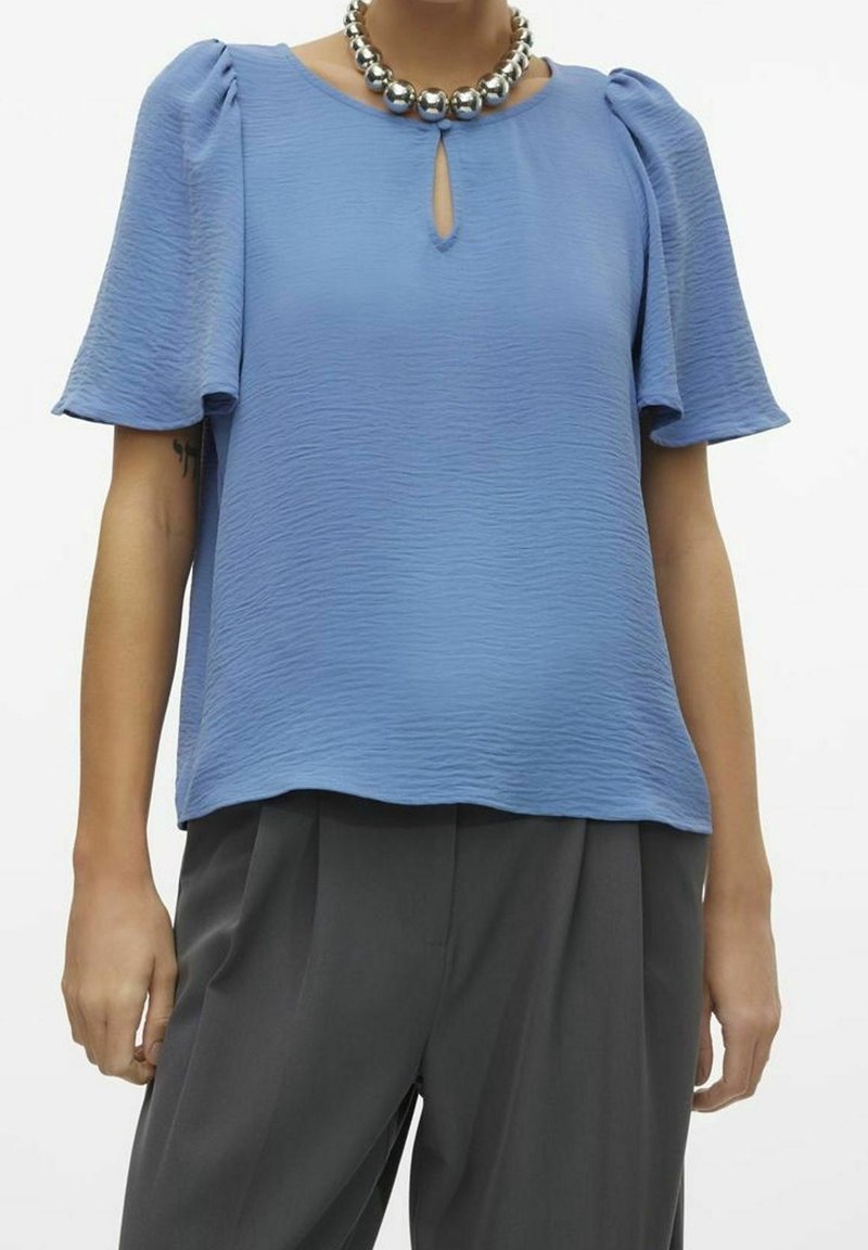 Blusa azzurro chiaro con tessuto testurizzato, scollatura a chiave e maniche corte a sbuffo. Abbinata a pantaloni grigi e una collana metallica rotonda.