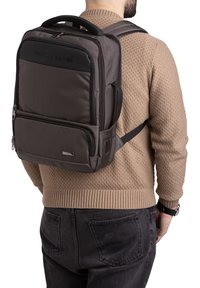 Zaino grigio scuro in nylon con schienale imbottito, spallacci regolabili e una tasca frontale con zip. Presenta un logo e dettagli metallici.