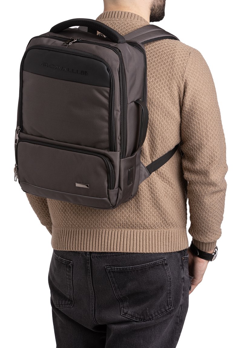 Zaino grigio scuro in nylon con schienale imbottito, spallacci regolabili e una tasca frontale con zip. Presenta un logo e dettagli metallici.