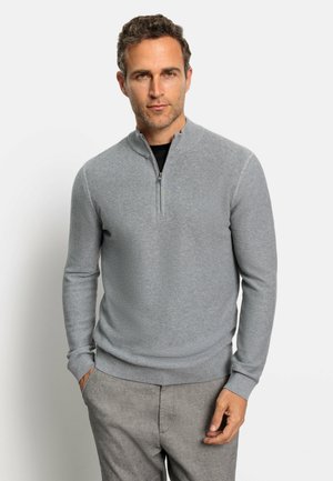 Hellgrauer, gerippter Strickpullover mit Reißverschluss am Hals. Mit langen Ärmeln und enganliegender Passform. Kombiniert mit hellgrauen Hosen.