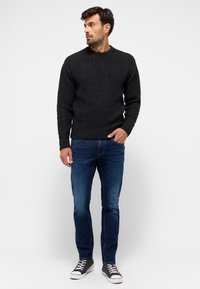 Mann steht in einem strukturierten schwarzen Pullover, dunklen blauen Jeans und schwarzen High-Top-Sneakern, schaut nach links vor einem weißen Hintergrund.