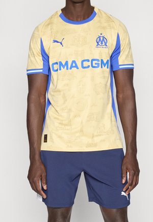 Mężczyzna w żółto-niebieskiej koszulce piłkarskiej Olympique de Marseille z logo CMA CGM i granatowych spodenkach Puma na białym tle.