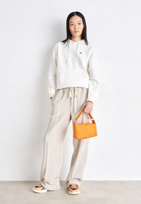 Felpa bianca con tasca frontale, pantaloni beige a gamba larga e strutturati, borsa arancione e sneakers con dettagli arancioni e navy.