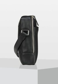 Sac bandoulière en cuir noir vu de profil avec une fermeture éclair argentée visible et une boucle réglable sur la sangle, posé sur une surface grise.
