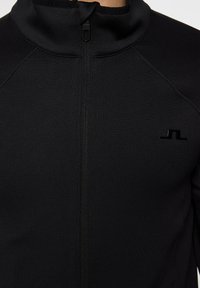 Svart zip-up jacka med högkrage, tillverkad av ett glatt och stretchy tyg, samt en liten logotyp på vänster bröst.