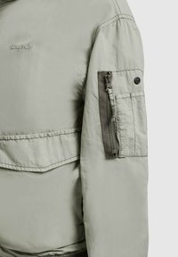 Manche de veste beige clair montrant une poche utilitaire zippée et un rabat boutonné, avec un texte de marque cousu sur la partie supérieure de la poitrine.