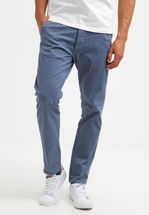 Man wearing blue slim-fit pants, white sneakers, and a white shirt with hands in pockets, standing on a white background.