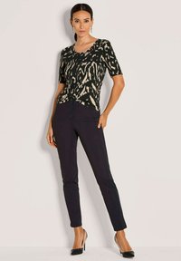 Vrouw die tegen een muur staat, gekleed in een zwart en beige gemusterd shirt met korte mouwen, zwarte broeken en zwarte hoge hakken.