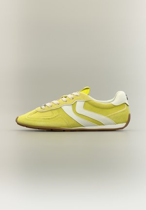 Sneaker in suede giallo con strisce laterali bianche, lacci bianchi e suola in gomma marrone, mostrato di profilo destro su sfondo neutro.