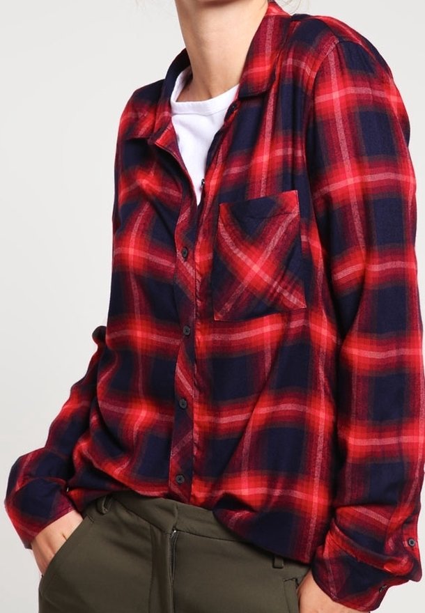 Persona che indossa una camicia a quadri rossa e blu navy sopra una maglietta bianca, con le mani parzialmente nelle tasche di pantaloni verde oliva.