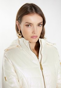 Geacă albă pufoasă cu un finisaj lucios, fermoar auriu și glugă cu șnur. Asortată cu cercei mari în formă de inel aurii.