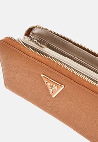 Tasche aus taupefarbenem Material mit goldfarbenen Beschlägen, ausgestattet mit einem dreieckigen Logoemblem. Der Reißverschluss öffnet sich zu einem hellen Innenraum.