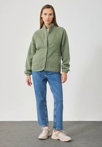 Roheka voodriga roheline fliisjope, millel on tekstuurse viimistlusega, nööpidega kinnitusega ja kaks eesmist taskut, kombineeritud helesiniste teksade ja beežide tossudega.