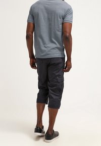 Homme debout de dos, portant une chemise à manches courtes bleu clair, un pantalon cargo capri noir et des baskets noires à semelles blanches.