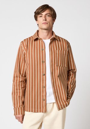 Homme portant une chemise boutonnée rayée verticale marron et blanche sur un t-shirt blanc et un pantalon beige, debout devant un fond uni.