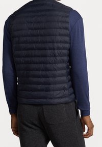 Marinblå pufferväst med quiltat mönster, med hög krage och ärmlös stil, bärs över en långärmad blå skjorta.