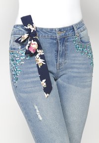 Jeans skinny azules con bordado floral en verde azulado y blanco, un lazo en color náutico como acento, y detalle desgastado en el muslo.