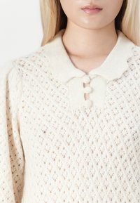 Frau trägt einen cremefarbenen, gestrickten Pullover mit einem gewellten Kragen und drei mit Stoff bezogenen Knöpfen am Ausschnitt.