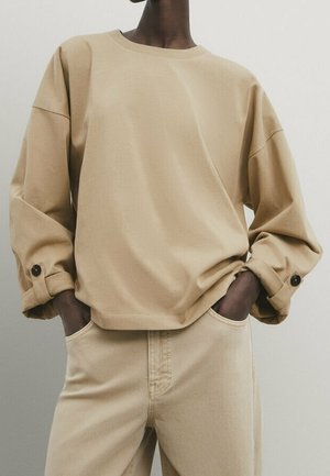 Personne portant une chemise ample beige à manches longues avec des poignets boutonnés et un pantalon beige, les mains dans les poches, devant un fond neutre uni.