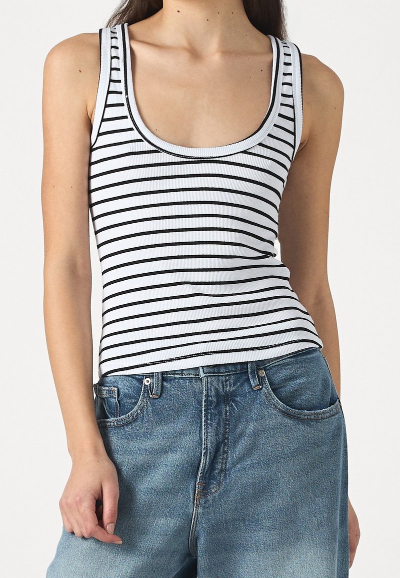 Femme portant un débardeur blanc sans manches à rayures horizontales noires et un jean bleu taille haute, les bras détendus le long du corps.