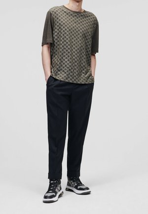 Oliwkowo-zielona koszulka z krótkim rękawem z geometrycznym wzorem, zestawiona z ciemnymi, dopasowanymi spodniami i czarno-białymi, wzorzystymi sneakersami.