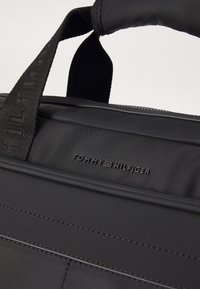 Sac en tissu noir avec une surface texturée, doté d'une poignée supérieure et d'un logo "TOMMY HILFIGER" embossé sur le devant.
