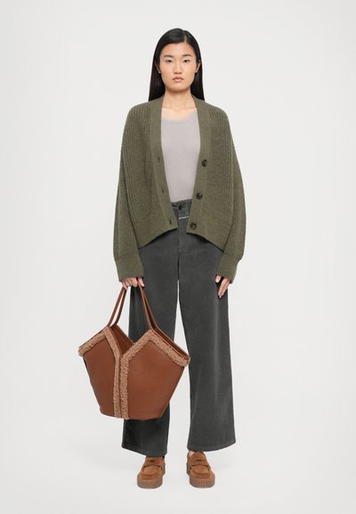 Woman wearing an olive knit cardigan, gray wide-leg pants, brown platform loafers, and holding a large brown tote bag with faux fur trim.