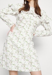 Vestido blanco de flores con patrones en rosa y verde, que presenta mangas con volantes y detalles fruncidos, fabricado en tela ligera.