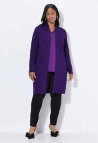 Frau in einem langen lila Cardigan über einem lila Oberteil, kombiniert mit schwarzen Hosen und schwarzen High Heels mit Schleifen, stehend vor einem weißen Hintergrund.