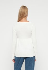 Bílý svetr s dlouhými rukávy, ribbovanou strukturou a kabelkovým vzorem v pase. Stylizováno s volnými modrými džíny.