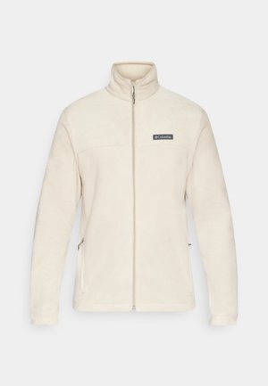 Veste en polaire beige avec un col montant, fermeture éclair intégrale et poches latérales. Elle présente un écusson logo noir sur la poitrine, une texture douce et des manches longues.