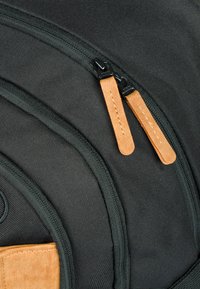 Zaino in tessuto nero con zip in pelle marrone chiaro, superficie texturizzata e cuciture curve. Presenta una tasca laterale con una finitura simile alla suede.
