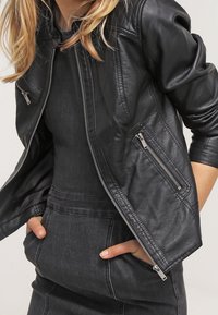 Žena v černé kožené bundě přes černé upnuté šaty, s rukama v kapsách šatů, na jednobarevném pozadí.