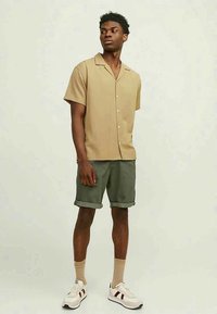 Beige shirt met korte mouwen, in een relaxte pasvorm met kraag, gecombineerd met groene shorts. Het model draagt witte sneakers met bordeauxrode accenten en sokken.
