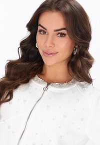 Valkoinen bomber-takki vetoketjulla, koristeltu helmipintaisella tekstuurilla ja kimaltelevalla jalokivikauluksella. Sileä kangas, istuva malli.