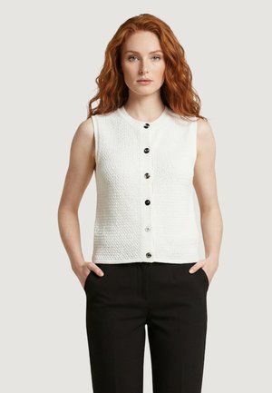 Donna con capelli rossi e mossi indossa una camicetta bianca senza maniche con bottoni e pantaloni neri, con le mani nelle tasche, sfondo neutro.