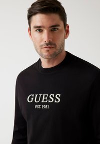 Felpa nera in tessuto con collo rotondo, caratterizzata da un ricamo del logo "GUESS" e "EST. 1981" in bianco a contrasto.