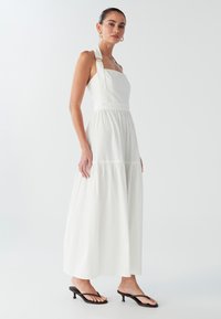 Vestido largo blanco con un corsé ajustado y falda en capas. Presenta un acento metálico en las tirantes y una textura suave. Se lleva con sandalias negras.