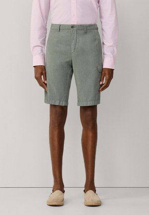 Homme portant un short cargo vert, une chemise à manches longues rose clair boutonnée et des espadrilles beige à enfiler, debout devant un fond uni.
