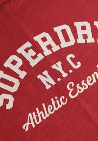 T-shirt en coton rouge avec le texte graphique blanc "SUPERDRY NYC Athletic Essentials" dans des polices en gras et en script. Texture douce, design décontracté.